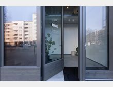 Geöffnete Glastüren zu einem modernen Gebäudeeingang mit minimalistischer Inneneinrichtung, darunter eine weiße Wand, ein Pflanzentopf und ein Hocker. Im Hintergrund sind durch die Scheiben mehrstöckige Wohngebäude und eine urbane Straße sichtbar.