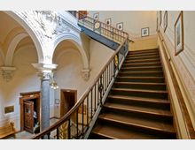 Historisches Treppenhaus mit breiter, hölzerner Treppe und kunstvoll verzierten Geländern, flankiert von Säulen mit dekorativen Kapitellen und Rundbögen. An den Wänden hängen Gemälde, und die Decke sowie die Wände sind mit Stuckelementen verziert.