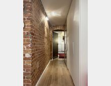 Ein schmaler Flur mit einer Wand aus rotem Sichtmauerwerk auf der linken Seite und weißen Einbauschränken rechts. Der Boden besteht aus hellem Holz, und im Hintergrund führt der Flur durch einen Backsteinbogen zu einer weiteren Tür und einem Raum mit weiß gestrichenen Wänden und einem roten Teppich.