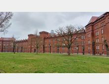 Ziegelrotes Gebäude im neugotischen Stil mit zahlreichen Spitzbogenfenstern und Türmen, umgeben von einem Rasen und vereinzelten Bäumen. Der Himmel ist bewölkt, und im Hintergrund ist eine moderne, gläserne Dachkonstruktion sichtbar.