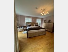 Ein modern eingerichtetes Schlafzimmer mit einem großen, gepolsterten Bett in der Mitte, flankiert von Nachttischen. Der Raum hat einen Holzboden, pastellfarbene Wände, zwei Fenster mit weißen Rollos und eine zentrale Deckenleuchte mit mehreren Glühbirnen.