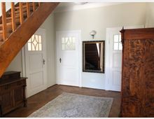 Wohnbereich mit Holztreppe, zwei weißen Zimmertüren mit verglasten Elementen, einem großen Wandspiegel und antiken Holzmöbeln auf einem Parkettboden. Ein heller Teppich bedeckt teilweise den Boden.