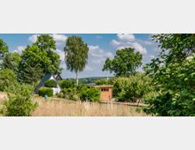 Gartenlandschaft mit kleinem weißen Haus in A-Form und Holzschuppen, umgeben von Bäumen, Sträuchern und Wiesen im Vordergrund. Im Hintergrund sind weitere Grünflächen und ein blauer Himmel mit wenigen Wolken zu sehen.