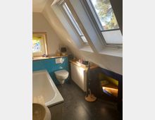 Badezimmer mit Dachschrägen und zwei großen Dachfenstern, die Tageslicht hereinlassen. Links befinden sich eine Badewanne und blaue Fliesen in Fischgrätmuster an der Wand, rechts eine Toilette, ein Heizkörper und ein Regal mit Ablage.