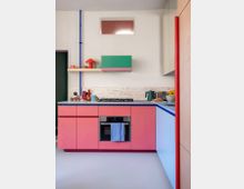 Kleine moderne Küche mit bunten Elementen, darunter rosafarbene Schränke, eine grüne Hängeschranktür und eine Arbeitsplatte aus hellem Stein. An der Wand befindet sich ein schlichtes Regal mit Dekorationsobjekten, und eine blaue senkrechte Leitung hebt sich von der weißen Wand ab.
