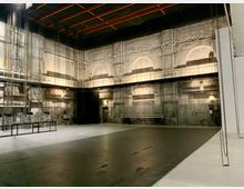 Ein leerer, industrieller Bühnenraum mit hohen grauen Wänden und sichtbarer Struktur. Über dem Boden verlaufen Seile und Techniksysteme, die von der Decke hängen. Eine erhöhte Galerie mit Geländer verläuft entlang der Rückwand.