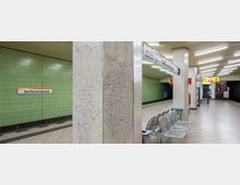 U-Bahn-Station Kurfürstendamm mit grünen gefliesten Wänden, Säulen mit kleinen Mosaikfliesen und einer Sitzreihe aus Metallbänken. Deckenleuchten beleuchten den Bereich, und Hinweisschilder sind über Fahrtrichtungen und Notrufinformationen angebracht.