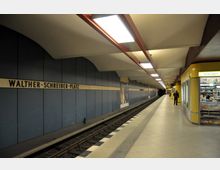 U-Bahnhof Walther-Schreiber-Platz in Berlin mit einer Bahnsteighalle, blauen Wandpaneelen, Deckenleuchten und einem Zeitungskiosk auf der rechten Seite. Die Bahngleise verlaufen entlang des Bahnsteigs, wo eine Person in der Ferne geht.