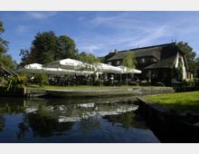 Ein Café mit Reetdachhaus und Terrasse, umgeben von grünen Bäumen und Rasen, liegt direkt an einem kleinen Wasserkanal, auf dem ein Holzboot vertäut ist. Auf der Terrasse stehen Sonnenschirme und Tische mit Gästen.