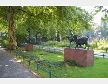 Kleiner Park mit einem Weg aus Kopfsteinpflaster, Rasenflächen und einer großen Platane im Vordergrund. Zwei Bronzeskulpturen auf roten Backsteinsockeln, darunter eine Figur mit einem Ziegenbock, befinden sich in einem umzäunten Bereich, umgeben von Blumenbeeten und Büschen.