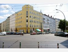 Straßenszene in einem städtischen Viertel mit mehrstöckigen Wohngebäuden, darunter ein gelbes Eckhaus mit Geschäften im Erdgeschoss und mehreren geparkten Autos entlang der Straße. Oberleitungen für Straßenbahnen und ein Fahrradfahrer sind im Vordergrund zu sehen.