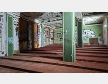 Innenraum eines verlassenen Gebäudes mit breiten, stufenartigen Podesten, grün-weiß gestrichenen Säulen und stark abblätternder Decken- und Wandfarbe. Wände und zugedeckte Fenster sind mit Graffiti bedeckt, im Hintergrund sind alte Holzrahmenfenster und eine kleine Öffnung im Boden zu sehen.