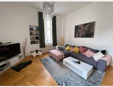 Wohnzimmer mit heller Wandfarbe und Parkettboden, einer grauen Sofaecke mit bunten Kissen, einem weißen Couchtisch und einem Teppich mit Muster. An der Wand hängt ein Bild eines Elefanten, und ein Fenster mit langen Vorhängen sorgt für Tageslicht. Links steht ein Fernseher auf einem weißen TV-Schrank, und in einer Wandnische sind Bücher und Dekorationen platziert.