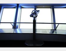 Innenraum eines Aussichtsturms mit großen, schrägen Fensterfronten und einem fest installierten Fernglas auf einem Metallständer. Die Umgebung wirkt durch das einfallende Licht modern und minimalistisch.