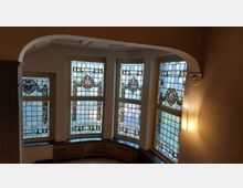 Eine Innenecke mit bogenförmigem Durchgang und fünf großen Fenstern mit bunten Bleiglasornamenten, die Wappen und Verzierungen zeigen. Rechts ist eine Wandlampe sowie ein dekoratives Gesims mit einem kleinen Skulpturmotiv erkennbar.