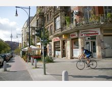 Straßenszene in einer urbanen Umgebung mit mehrstöckigen Altbauten, einem Restaurant mit Straßenterrasse und einem Vorbeifahrenden auf einem Fahrrad. Im Hintergrund ist der Berliner Fernsehturm sichtbar, die Straße ist flankiert von geparkten Autos und Bäumen.