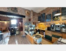 Gemütliches Café mit rustikalem Backstein-Interieur, einem Tresen mit Holzmöbeln und Kaffeemaschinen sowie einer Auslage mit Geschirr und Speisen. Im Hintergrund befinden sich weitere Sitzbereiche mit Holztischen und Stühlen.