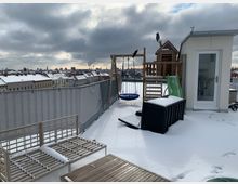 Schneebedeckte Dachterrasse mit Blick auf eine Stadtlandschaft. Auf der Terrasse befinden sich Gartenmöbel, ein kleiner Kinderspielplatz mit Schaukel, Kletterturm und Rutsche sowie ein Zugang zu einem weißen Anbau mit Fenstertür.