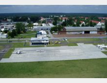 Kleiner Regionalflughafen mit einem Hauptgebäude, einer asphaltieren Start- und Landefläche sowie zwei geparkten Kleinflugzeugen. Im Hintergrund sind Lagerhallen, kleinere Gebäude und Bäume zu sehen.