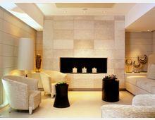 Moderner, eleganter Wohnbereich mit hellen Wänden aus Naturstein, großem rechteckigem Kamin mit drei brennenden Kerzen, beigen Polstermöbeln und schwarzen Beistelltischen. Dezente Beleuchtung und dekorative Elemente wie goldene Ornamente komplettieren die Einrichtung.