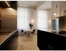 Moderne Küchen- und Essecke mit dunklen, minimalistischen Einbauschränken und einer großen Kücheninsel mit Kochfeld. Vor zwei großen Fenstern mit weißen, bodenlangen Vorhängen steht ein runder Tisch mit Stühlen auf hellem Holzboden.