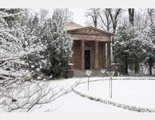 Schneebedecktes Mausoleum mit Säulenfassade, umgeben von Bäumen und einem gepflegten Gartenweg. Die winterliche Landschaft ist detailreich mit schneebedeckten Ästen und Hecken gestaltet.