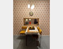 Ein retro-inspiriertes Wohnzimmer mit gemusterten Tapeten in Braun- und Beigetönen, einem rechteckigen Tisch mit einer bestickten Tischdecke, umgeben von senfgelben Stühlen, und einer gelben Schreibmaschine. Im Hintergrund ein Wanddurchbruch mit Regalen, die ein Radio, eine Thermoskanne und Dekorationsgegenstände enthalten, sowie eine Wanduhr oberhalb des Durchbruchs.