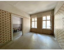 Leerer Innenraum in einem älteren Gebäude mit abgenutztem Boden und floral gemusterter Tapete an den Wänden. Zwei große Sprossenfenster lassen Tageslicht herein; links führt ein breiter Durchgang in einen Nebenraum.