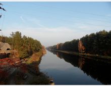 Blick auf einen geraden Fluss oder Kanal, umgeben von dichten, herbstlich gefärbten Nadel- und Laubbäumen. Links im Bild ist ein Gebäude mit Strohdach zu sehen, daneben ein kleiner Weg, auf dem Menschen spazieren gehen.