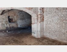 Ein alter, leerer Kellerraum mit grobem Mauerwerk aus Ziegelsteinen und einem gemauerten Rundbogen. Der Boden ist aus abgenutztem Pflaster, und die Wände zeigen Spuren von Verwitterung und abblätterndem Putz.