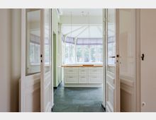 Blick durch geöffnete weiße Flügeltüren in eine helle Küche oder Veranda mit großflächigen Fenstern und gewölbtem Deckenbereich. Vor den Fenstern steht ein weißer Unterschrank mit Holzarbeitsplatte auf dunklem Steinfliesenboden.