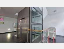 Innenbereich eines modernen Gebäudes mit einem Aufzug in Metallverkleidung und verglasten Wänden. Rechts sind Transportwagen neben einem Geländer mit Holzhandlauf zu sehen.