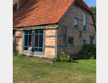 Fachwerkhaus mit roten Ziegelwänden, blauen Fensterrahmen und einem Dach aus roten Dachziegeln. Das Gebäude steht in einer grünen Gartenlandschaft mit niedrigen Sträuchern und einer Leiter vor der Fassade.