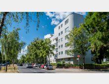 Mehrstöckiges Wohngebäude in moderner Bauweise mit weißen Fassaden und abgerundeter Ecke, umgeben von Bäumen und einer Straße, an der Autos parken. Im Vordergrund verläuft eine von Bäumen gesäumte Fahrbahn bei sonnigem Wetter.