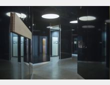 Ein moderner Ausstellungsraum mit glänzenden, schwarzen Wänden, mehreren Türen und beleuchteten Deckenlampen. Im Hintergrund sind helle Wände mit kleinen Informationsrahmen erkennbar.