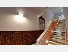 Treppenhaus mit moderner, hölzern-verkleideter Treppe und seitlicher Beleuchtung; an der Wand eine alte Zuganzeigetafel mit Uhr sowie eine Beschilderung. Die Wand ist teilweise mit dunkelbraunen Fliesen verkleidet.