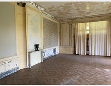 Ein leerer Raum in einem alten Gebäude mit maroden Wänden, teils verblasster Tapete und einer weißen Gardine, die vor einer Glastür hängt. Der Holzboden ist in Fischgrätmuster verlegt, und an der Wand steht ein kleiner Kaminsims mit einer schwarzen Vase.