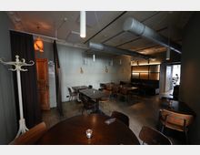 Innenraum eines modernen Restaurants mit mehreren dunklen Holztischen und Stühlen auf Betonboden. Sichtbare Lüftungsrohre und schlichte Hängelampen an der Decke sowie eine Sitzecke mit gepolsterter Bank und abgetrenntem Bereich im Hintergrund.