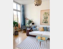 Helles Wohnzimmer mit großen Fenstern und blauen Vorhängen, einem modernen Sofa mit bunten Kissen, einem kleinen Couchtisch auf einem Zickzack-Teppich und mehreren Pflanzen. An der weißen Wand hängen ein großes Bild mit einer Palme und kleine Regale mit Dekorationen.