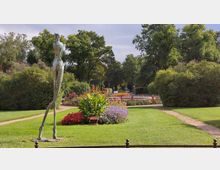 Ein gepflegter Park mit grünen Rasenflächen, blühenden Blumenbeeten und Gehwegen, im Vordergrund eine abstrakte Skulptur aus Metall. Im Hintergrund sind große Bäume und parkende Autos zu sehen.