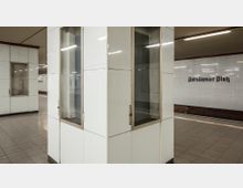 U-Bahn-Station mit weiß gefliesten Wänden und Boden aus grauen Fliesen, im Hintergrund die Beschriftung "Potsdamer Platz" an der Wand. Glasvitrinen und ein Abschnitt des Bahnsteigs sind sichtbar.