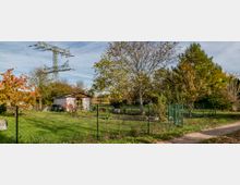 Kleingarten mit einer kleinen hölzernen Laube, umgeben von einem Zaun und zahlreichen Bäumen. Im Hintergrund stehen Strommasten, und auf der linken Seite ist eine grüne Wiese zu sehen.