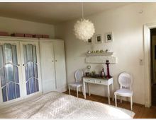 Helles Schlafzimmer mit weißer Einrichtung, darunter ein großer Schrank mit Glaseinsätzen, ein kleiner Schminktisch mit Kerzen und Dekorationen sowie zwei weiße Stühle. Eine dekorative Deckenlampe hängt in der Mitte, Boden aus Holz und Wände schlicht gehalten.