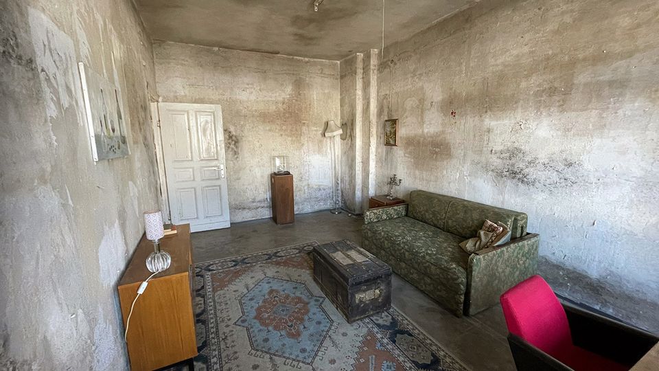 Schlichter, alt wirkender Innenraum mit hohen Decken, rau verputzten und teils fleckigen Wänden sowie einer weißen Tür an der Stirnseite. Im Raum stehen ein gemusterter Teppich, ein grünes Sofa, eine Truhe als Tisch und ein kleines Sideboard mit Lampen, beleuchtet von einer einfachen Deckenleuchte.