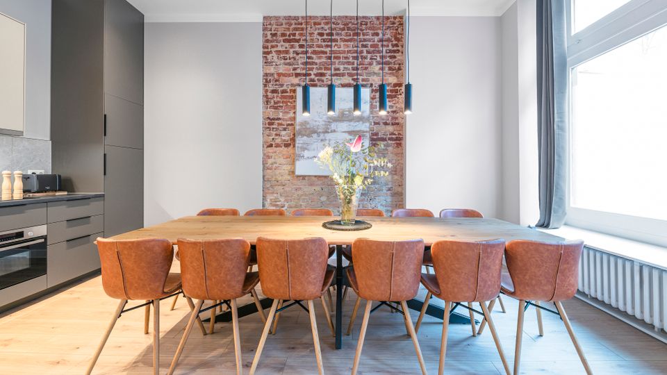 Moderner Essbereich mit großem Holztisch und mehreren braunen Lederstühlen, angrenzend an eine dunkle Einbauküche. Im Hintergrund eine freigelegte Ziegelwand mit Bild, darüber schlanke Pendelleuchten; rechts ein großes Fenster mit Vorhang und Heizkörper darunter.