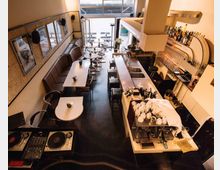 Ein modernes Café mit stilvoller Inneneinrichtung; links stehen Tische mit Stühlen und einer gepolsterten Sitzbank entlang der Wand, rechts befindet sich eine lange Theke mit Barhockern und einer Kaffeemaschine. Im Hintergrund sind große Fenster und ein Eingang zu sehen, die für viel Tageslicht sorgen.