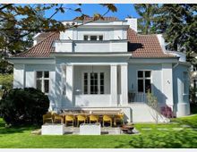 Weißes Einfamilienhaus im klassischen Stil mit roten Ziegeldach und symmetrischen Fenstern. Im Vordergrund eine Terrasse mit einem Holztisch, gelben Stühlen, Blumenkübeln und einer Feuerstelle, umgeben von einem gepflegten Garten.