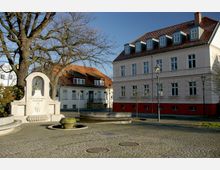 Ein kleiner Platz mit Kopfsteinpflaster, auf dem ein Denkmal mit einer Büste steht, umgeben von großen Bäumen, einer weißen Fassade mit roten Ziegeldächern und einem zweigeschossigen Gebäude mit hellem Putz und roten Sockeln. Im Vordergrund stehen runde Brunnenstrukturen, und Laternen säumen den Platz.