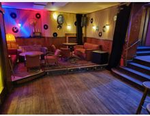 Gemütliche Lounge in einem Retro-Stil mit Holzboden, niedrigen Tischen, Sesseln und Sofas, dekoriert mit Schallplatten an den holzvertäfelten Wänden. Die gedämpfte Beleuchtung mit bunten Akzenten verleiht dem Raum eine warme Atmosphäre; eine kleine Treppe führt zu einem erhöhten Bereich.