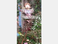 Eine Gartenszene mit einer dekorativen Büstenfigur aus Keramik auf einem Sockel, umgeben von grünen Ranken, verblühten Pflanzen und vereinzelten weißen Blüten. Im Hintergrund ist eine Mauer mit Efeu bewachsen.